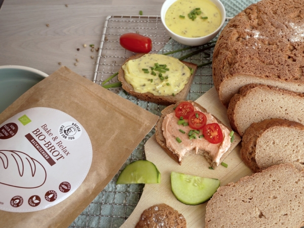 Böcker Brotbackmischung glutenfrei, in einer braunen Tüte auf Holztisch. Daneben auf einem Gitterrost ein angeschnittenes Brot sowie halbierte Brotstücke, die mit Streichcreme beschmiert sind und Stücke von Tomate, Gurke