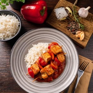 Ein grauer Steingutteller auf einer Holzoberfläche, darauf zwei Putenschschlik mit Reis und roter Sauce. Auf dem Tisch eine Reisschale, Paprika, Holzbrett mit Gewürzen und eine Gabel mit Serviette 