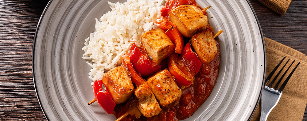 Zwei Putenschaschlik mit Paprika mit Reis und roter Sauce auf einem grauen Steingutteller, angerichtet auf Holztischoberfläche